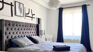 Quarto moderno com cabeceira estofada, cama grande com roupa de cama cinzenta, mesa de cabeceira e janela com cortinas azuis.