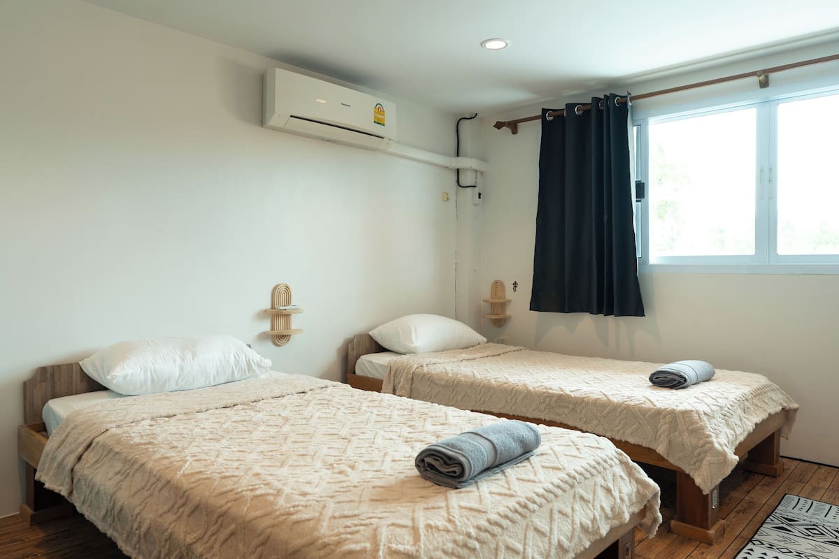 Two single beds with beige linens, a window with dark curtains, and an air conditioning unit on the wall.