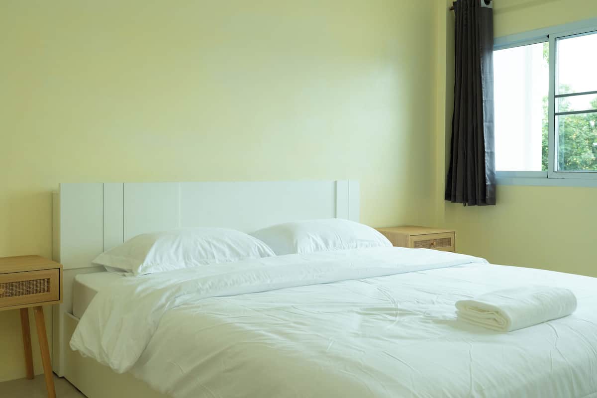 A neatly made bed with white linens and pillows, a bedside table, and a window with curtains.