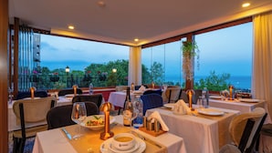 A restaurant with tables set for dining, featuring a view of the sea and a balcony with plants.