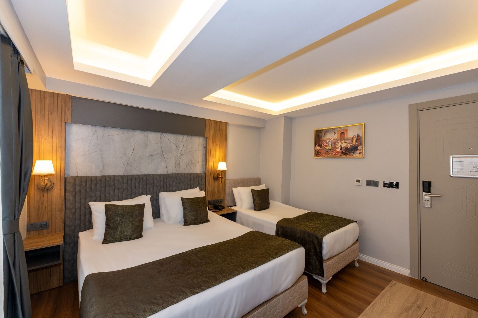 Quarto de hotel com duas camas, cabeceira de mármore, luminárias fixadas na parede, um quadro e uma porta com fechadura.