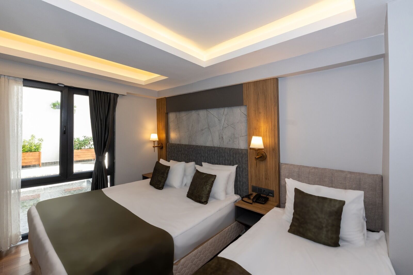 Ein Hotelzimmer mit zwei Betten, einem großen Fenster mit Vorhängen, einem hölzernen Kopfteil und einem Nachttisch mit Lampe.