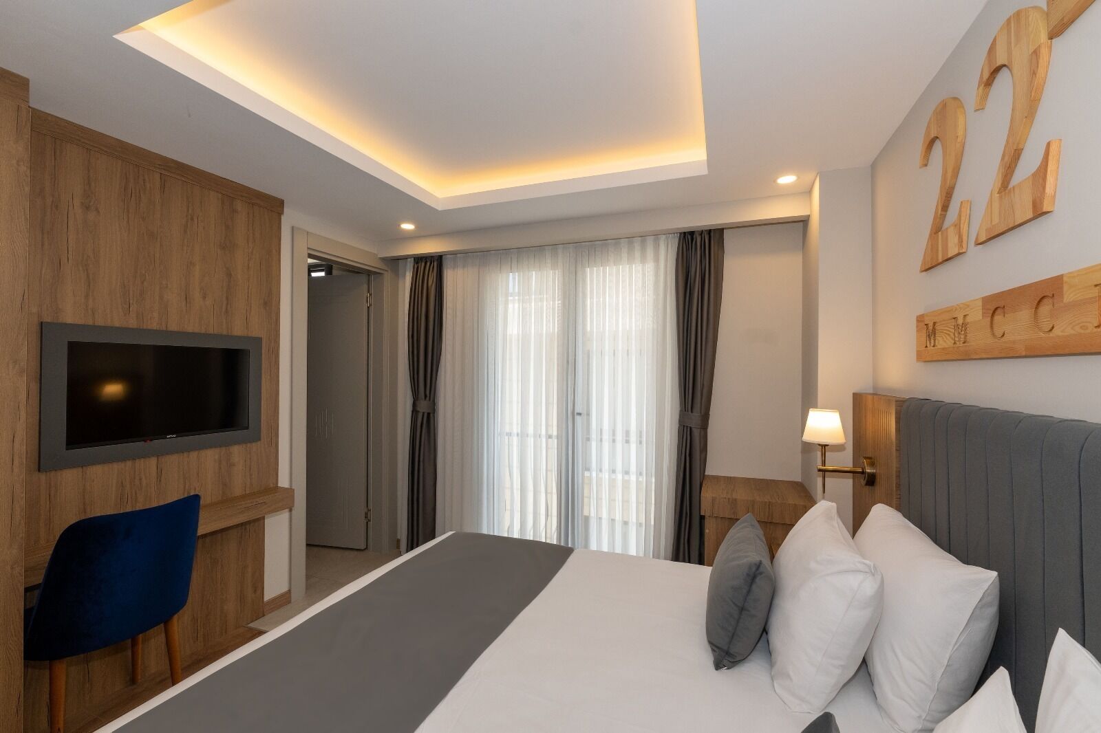 A hotel room with a large bed, a TV mounted on the wall, a desk with a chair, and a window with curtains.