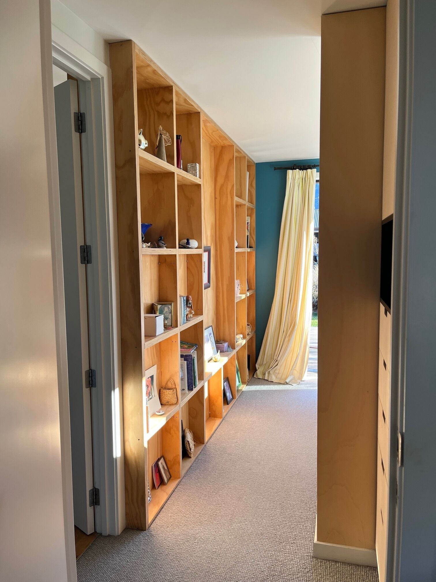 Un corridoio con una libreria in legno piena di libri e oggetti decorativi, che conduce a una stanza con un'ampia finestra e una tenda.