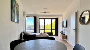Ein modernes Wohnzimmer mit einem runden Tisch, schwarzen Stühlen, einer Couch mit Kissen, einem an der Wand montierten Fernseher und einem großen Fenster mit Blick auf Gebäude und Bäume.