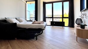 Ein modernes Schlafzimmer mit einem Bett, einem kleinen Nachttisch, einem Fernseher und großen Fenstern mit Blick auf die Stadt.