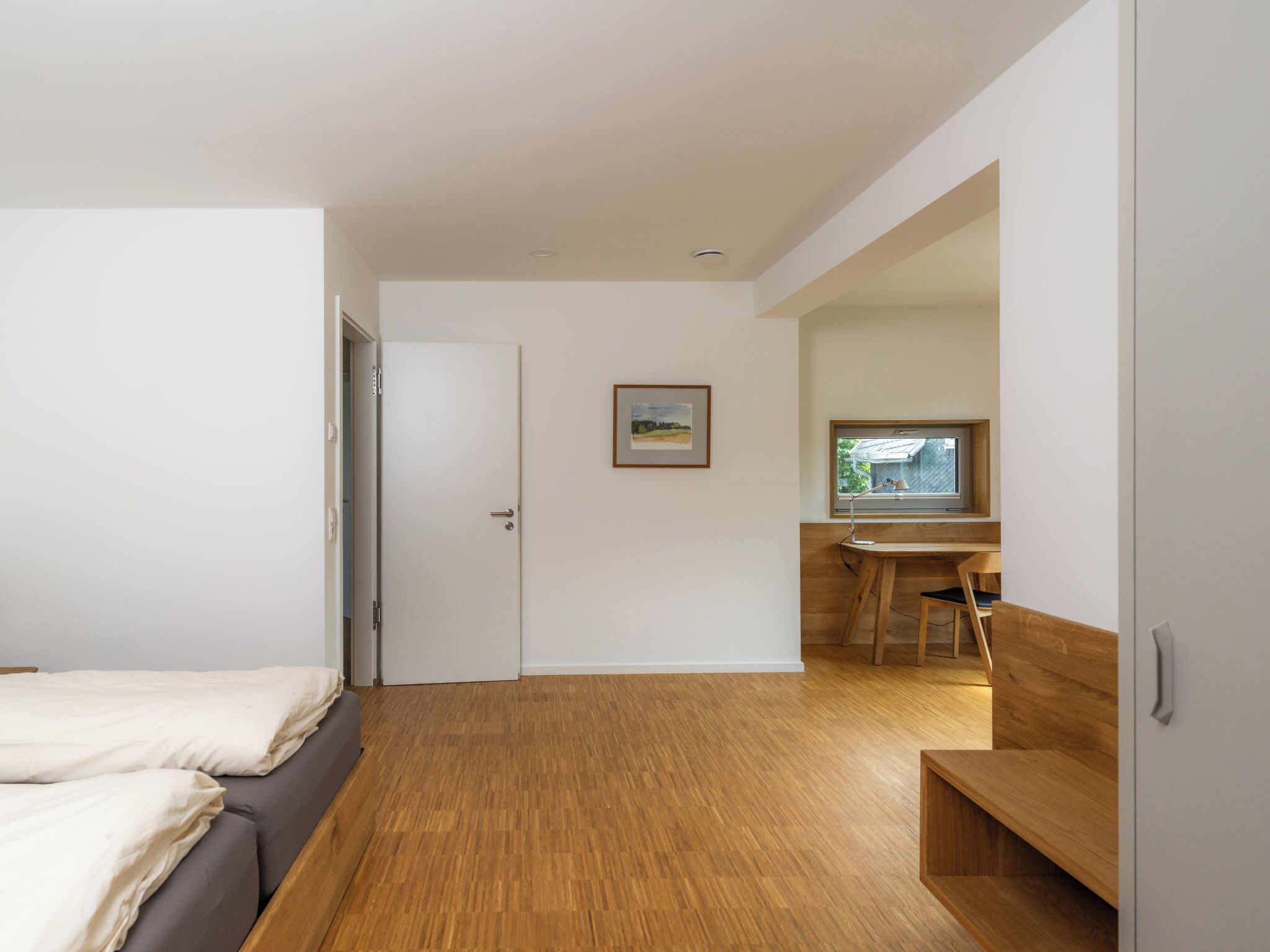 A modern, minimalist room with a wooden floor, a bed with white linens, a desk with a chair, and a window with a view of a building.
