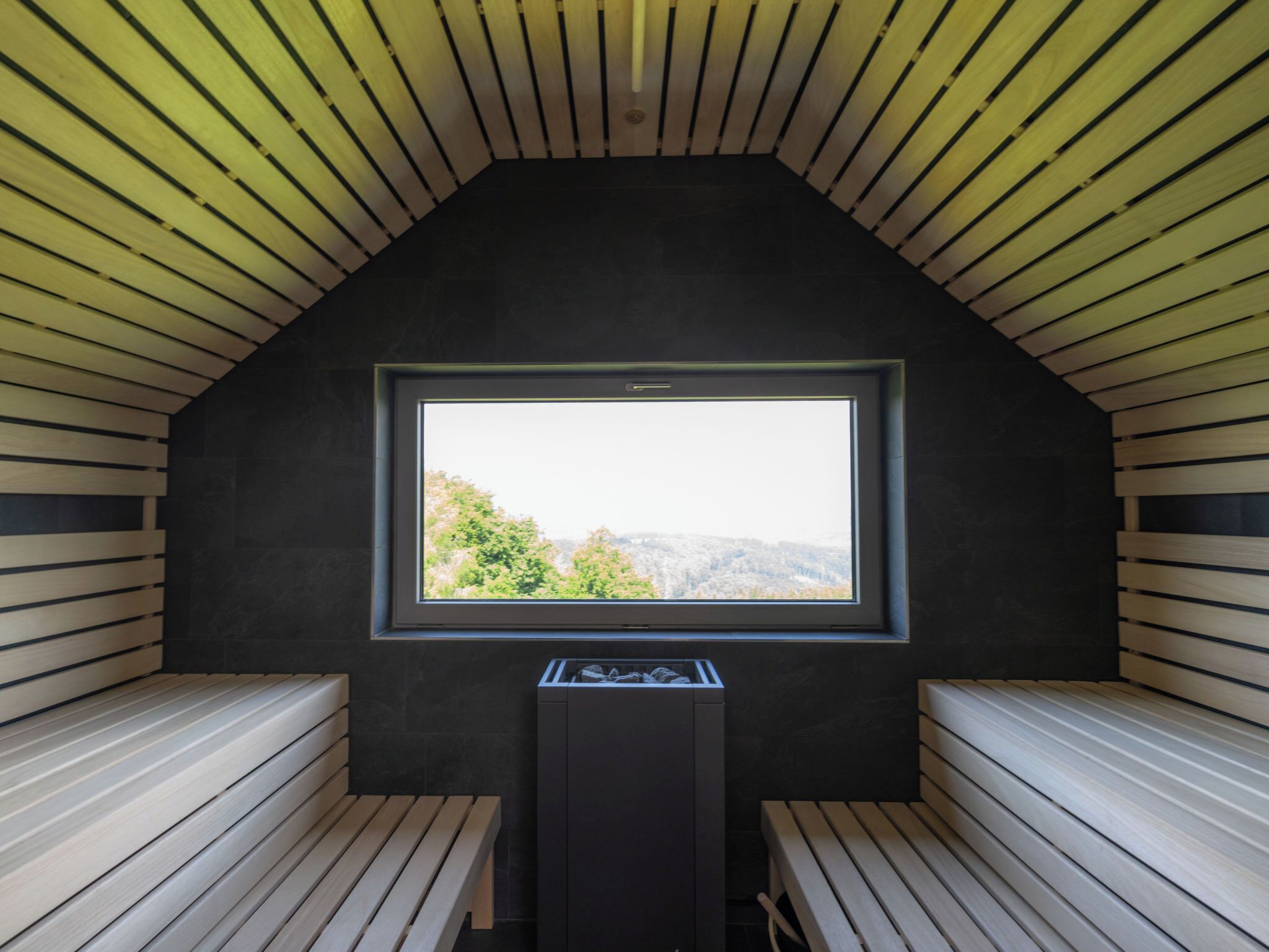 A room with a sloped ceiling, wooden benches, a central window, and a black cabinet.