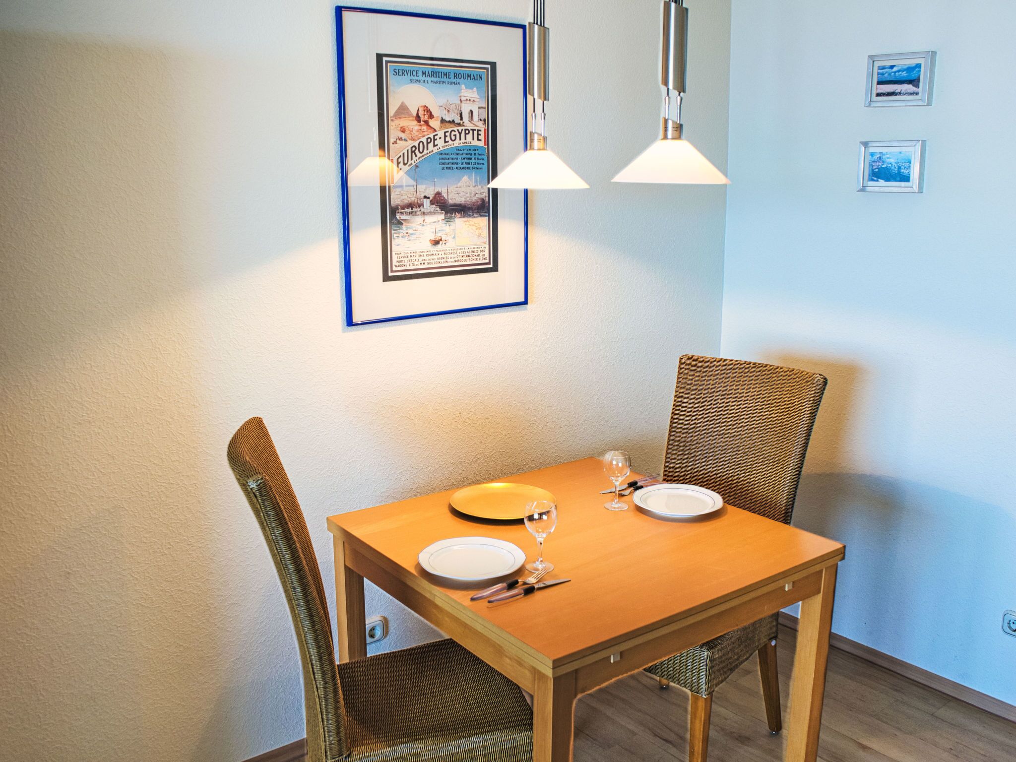 Een eetgedeelte met een houten tafel, twee rieten stoelen en een ingelijste poster aan de muur.