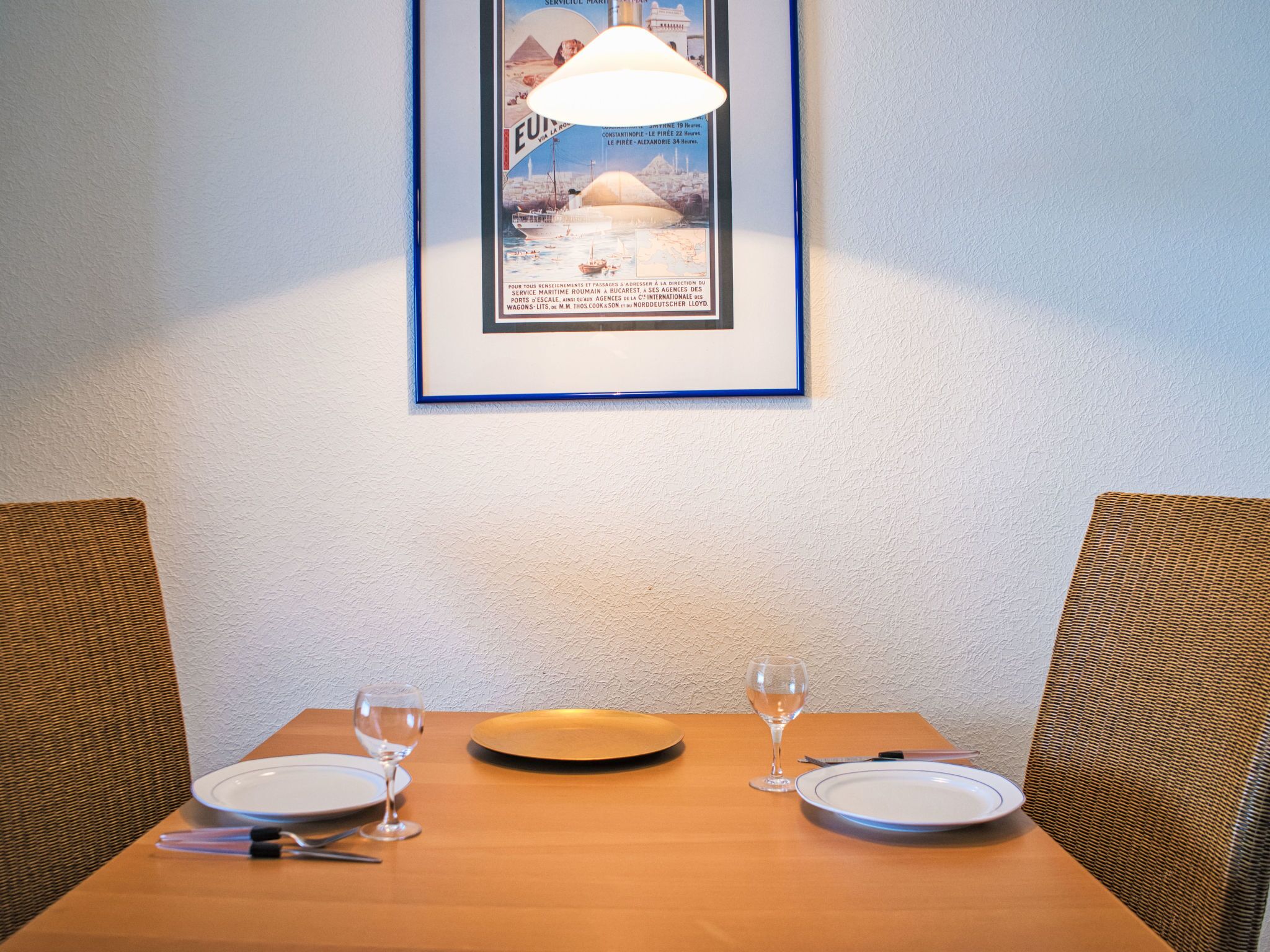 Et spisebord med to tallerkener, et vinglas og bestik, placeret foran en væg med struktur og et billede i ramme hængende over.