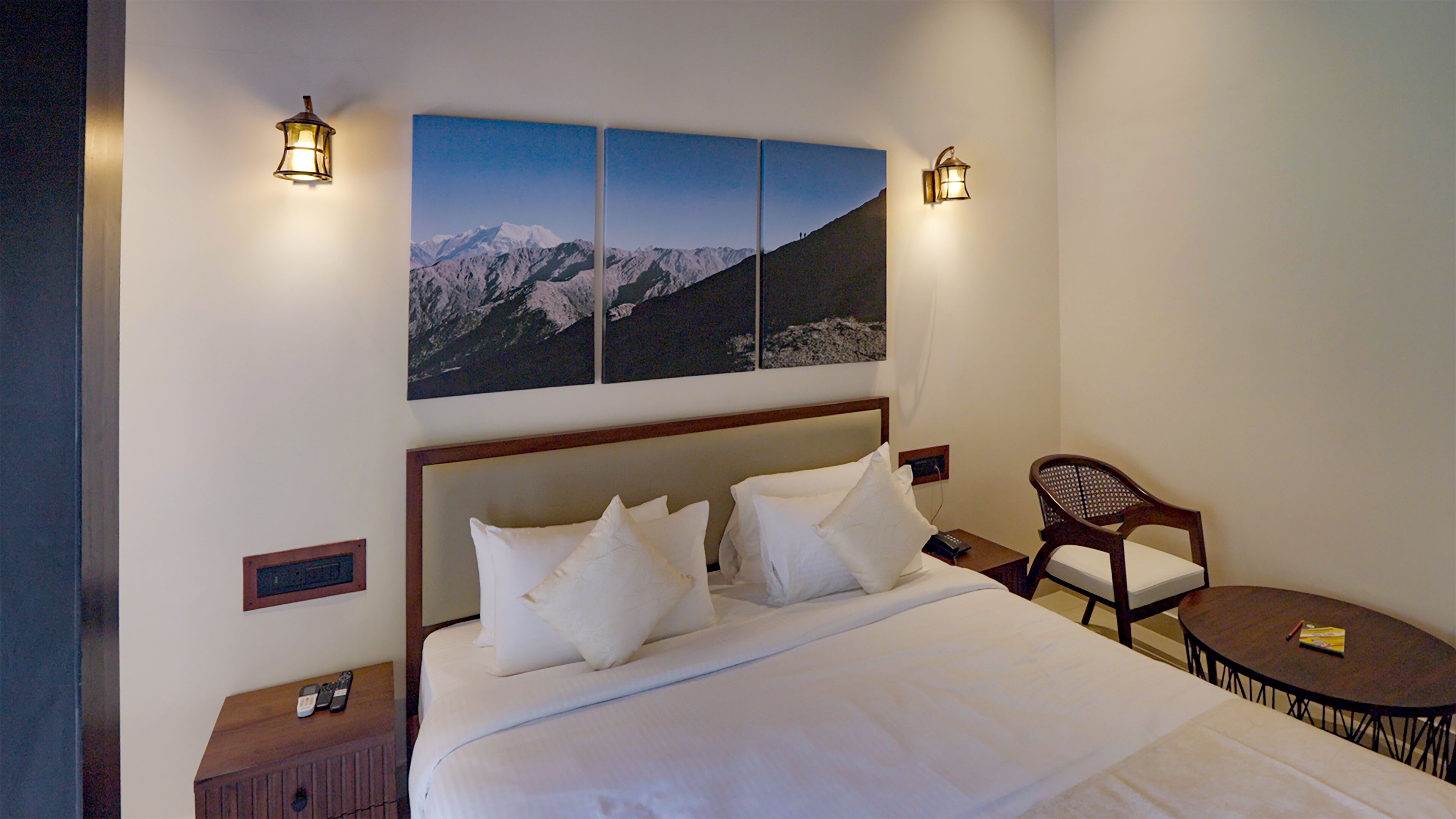 Camera d'albergo con un letto grande, due comodini, una sedia, un tavolino e un quadro appeso al muro raffigurante montagne.