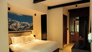 A bedroom with a bed, a chair, a desk, and a painting on the wall. - The Big Bridge (Rishikesh)