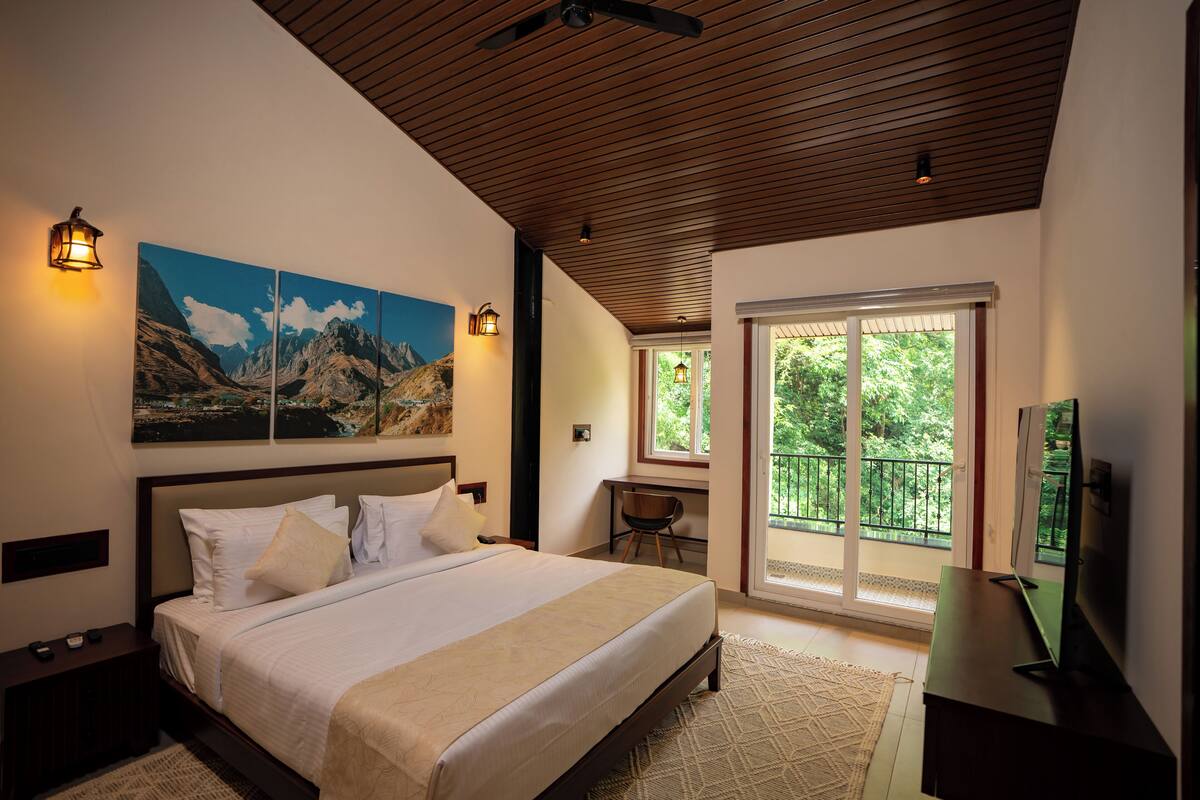A bedroom with a large bed, a desk, and a TV. There is a balcony with a view of trees.
