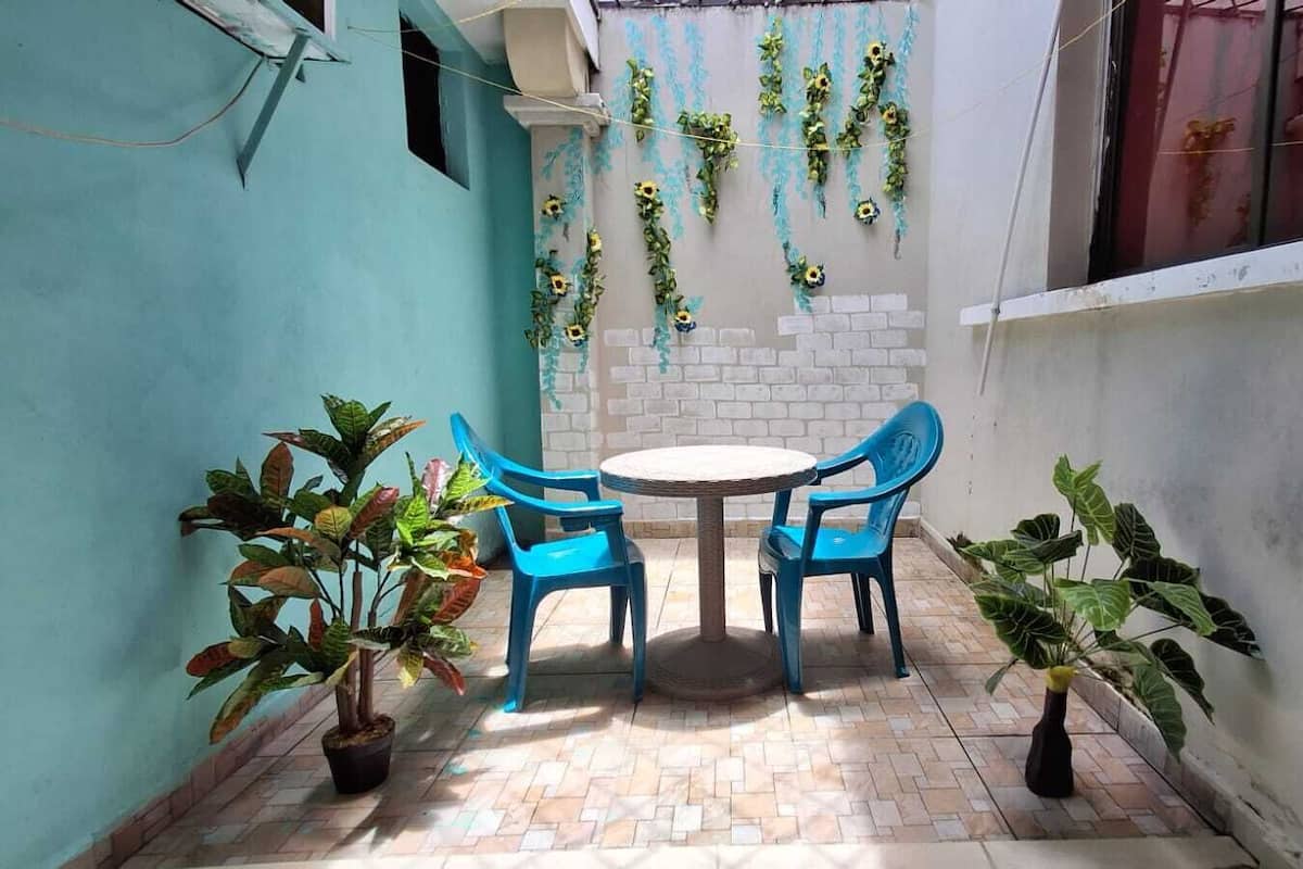 Um pequeno pátio com uma mesa redonda e quatro cadeiras azuis, rodeado por plantas em vasos.