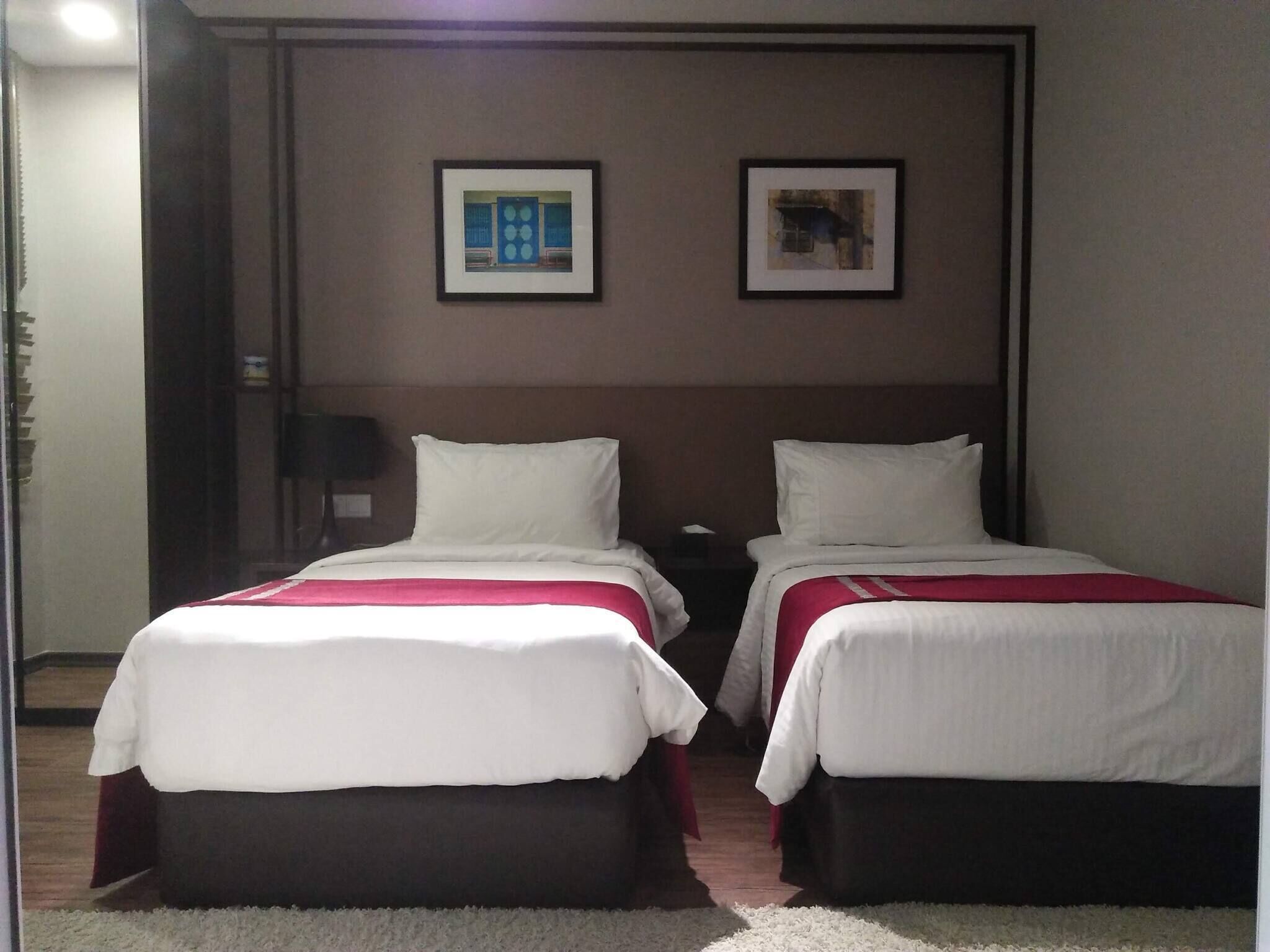 Habitación de hotel con dos camas, cada una con ropa de cama blanca y un detalle en rojo. Dos cuadros enmarcados decoran la pared sobre las camas.