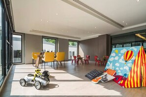 Un hall d’hôtel moderne avec de grandes fenêtres, des fauteuils jaunes et rouges, une petite voiture jouet et une décoration colorée en forme de tente.