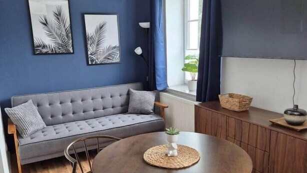 A modern living room with a grey sofa, a round wooden table, and a large window with blue curtains. - Studio 1 - Apartments at the castle (Oberschleißheim)
