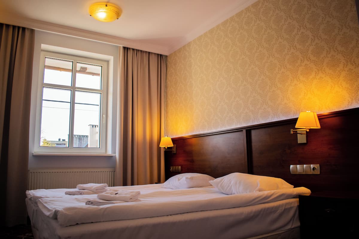A hotel room with a bed, two bedside lamps, a window with curtains, and a radiator.