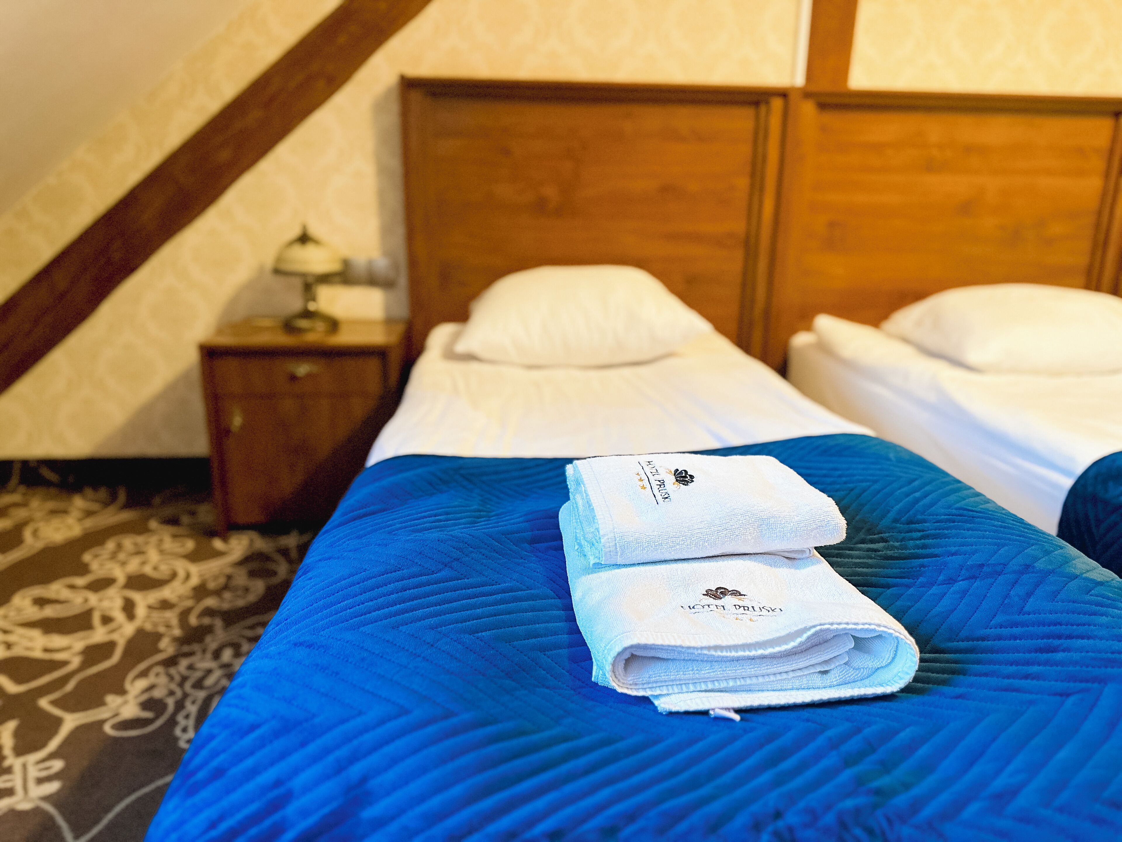 Habitación de hotel con cabecera de madera, dos camas con ropa blanca, colcha azul acolchada y una toalla doblada con un logotipo en la cama.