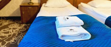 Habitación de hotel con cabecera de madera, dos camas con ropa blanca, colcha azul acolchada y una toalla doblada con un logotipo en la cama.