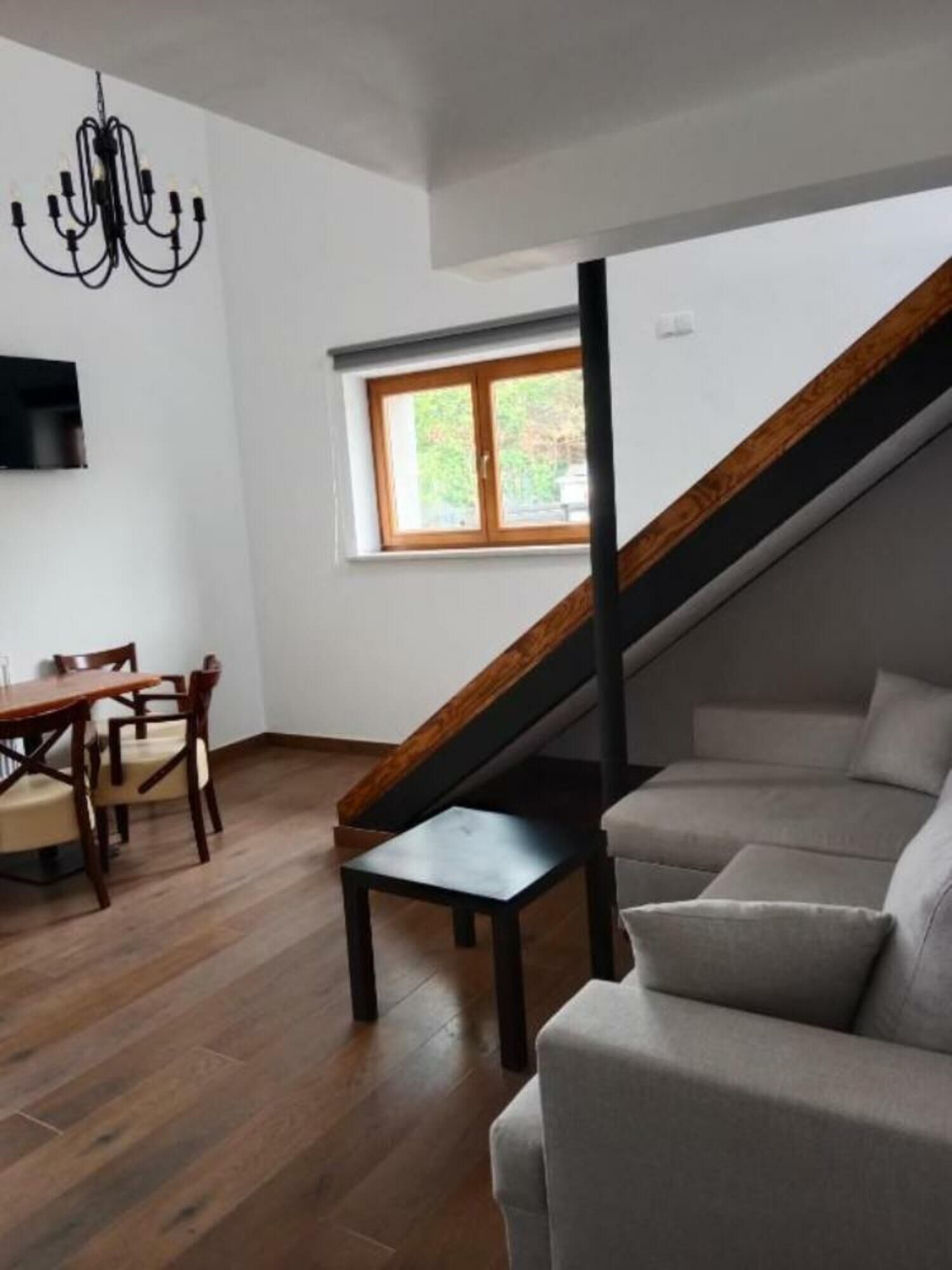 Una sala de estar moderna con una escalera de madera, un comedor con mesa y sillas, y un amplio ventanal que proporciona luz natural.