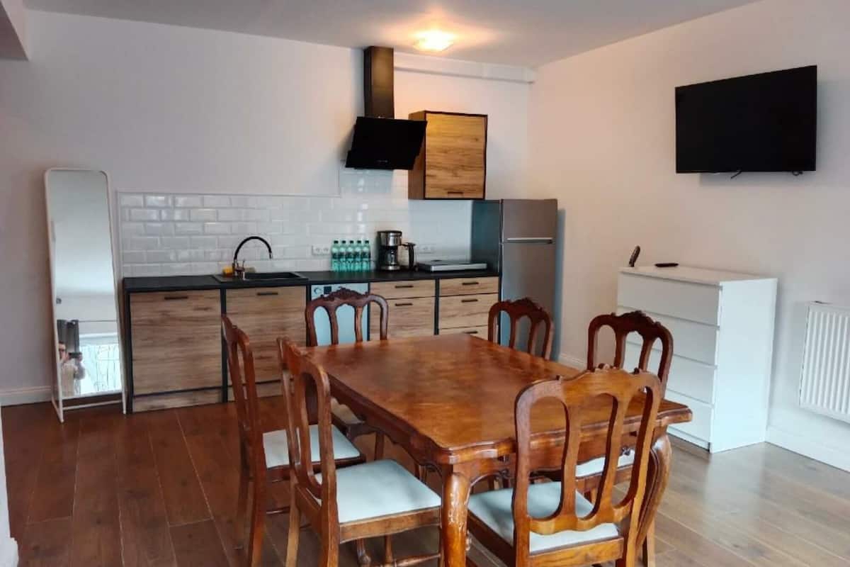 A modern kitchen with white subway tiles, wooden cabinets, and a built-in oven. There is a dining table with six chairs and a flat-screen TV mounted on the wall.