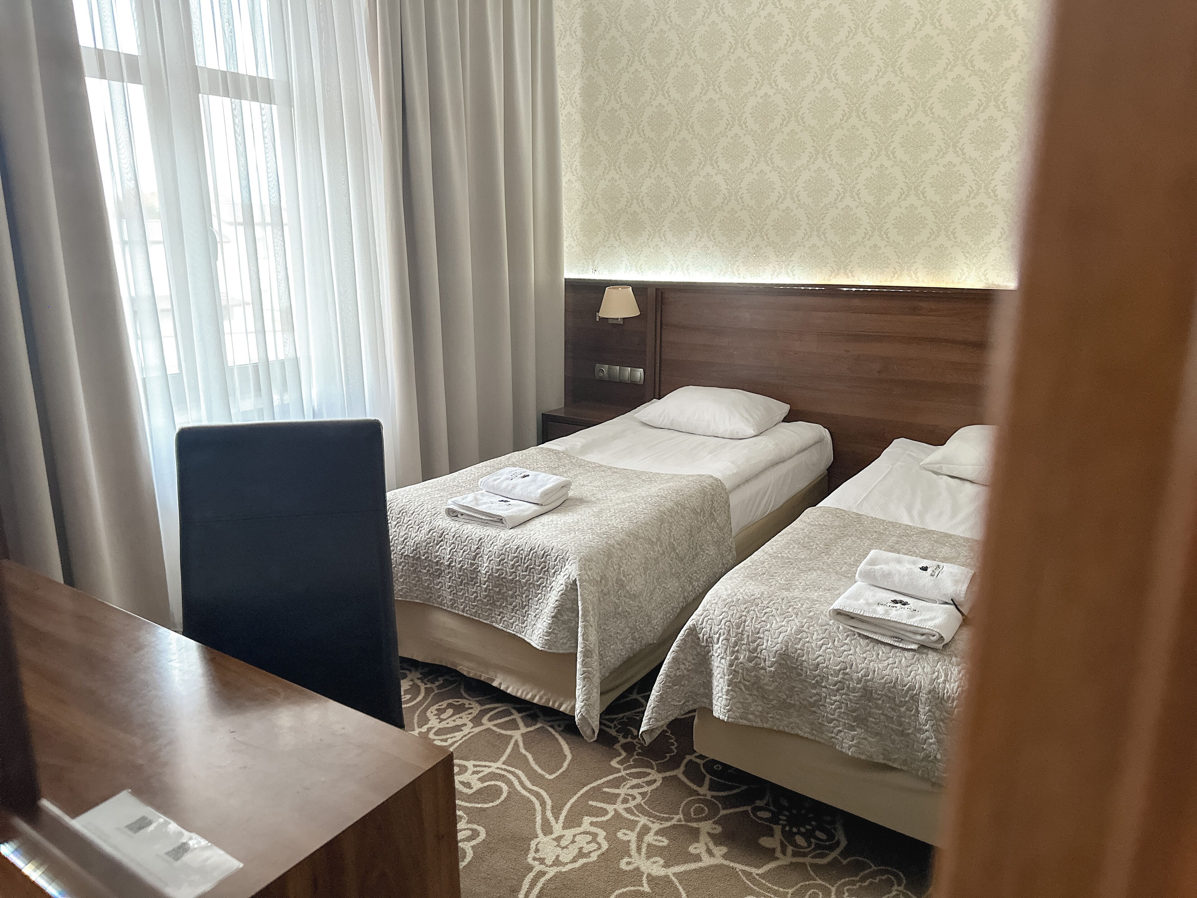 Habitación de hotel con dos camas, un escritorio, una silla, una mesita de noche con lámpara y una alfombra estampada.