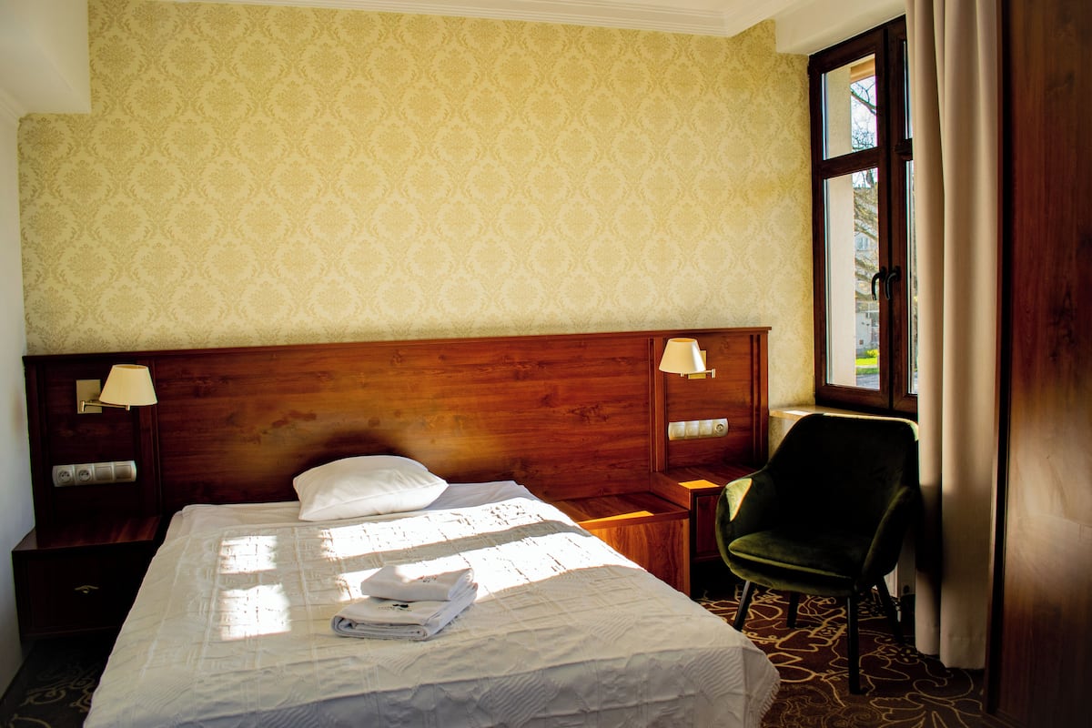 A hotel room with a large bed, two bedside lamps, a green chair, and a window with curtains.