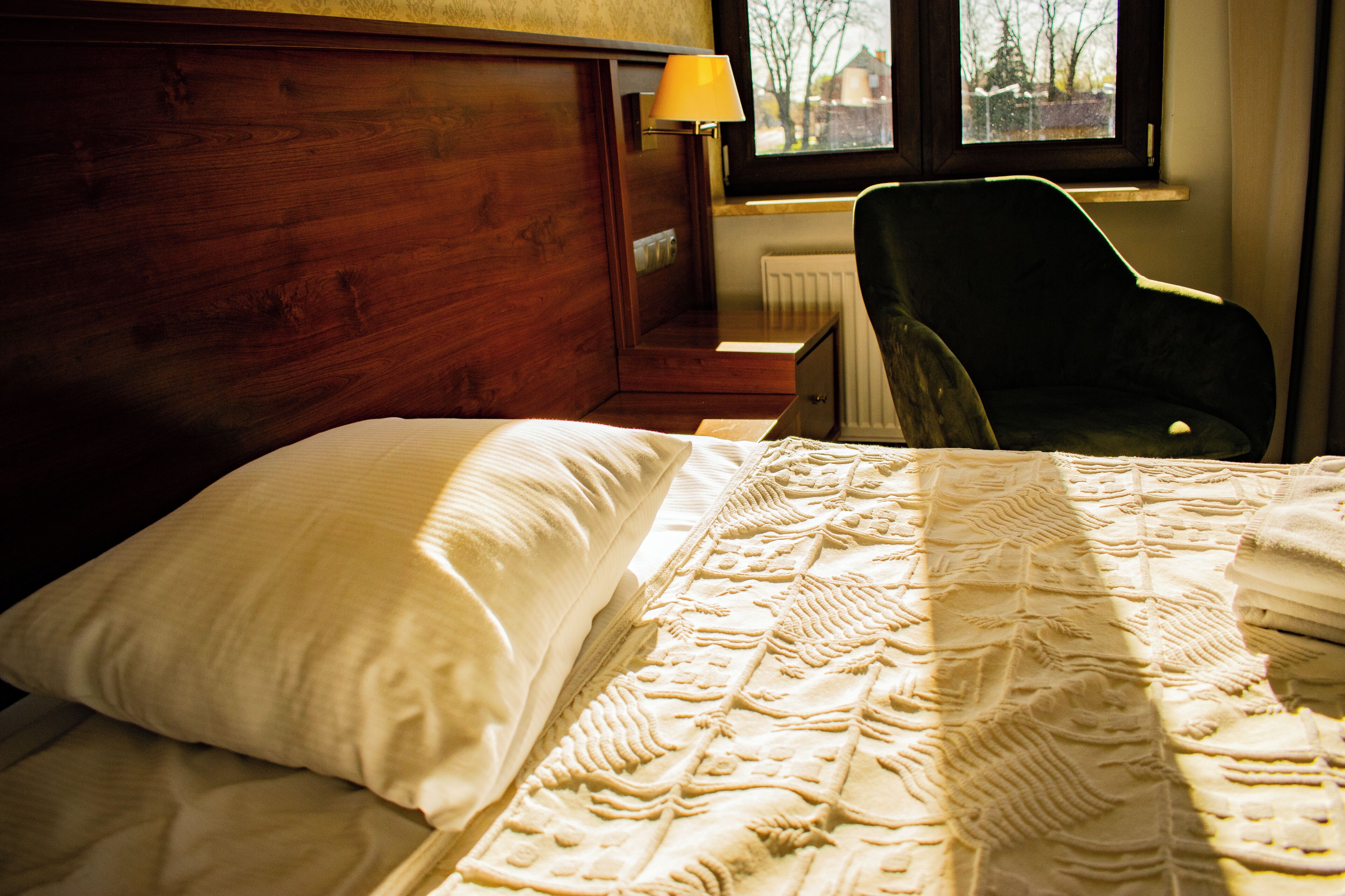 Une chambre d’hôtel avec une tête de lit en bois, un lit avec une literie blanche, une chaise verte et une fenêtre donnant sur des arbres.