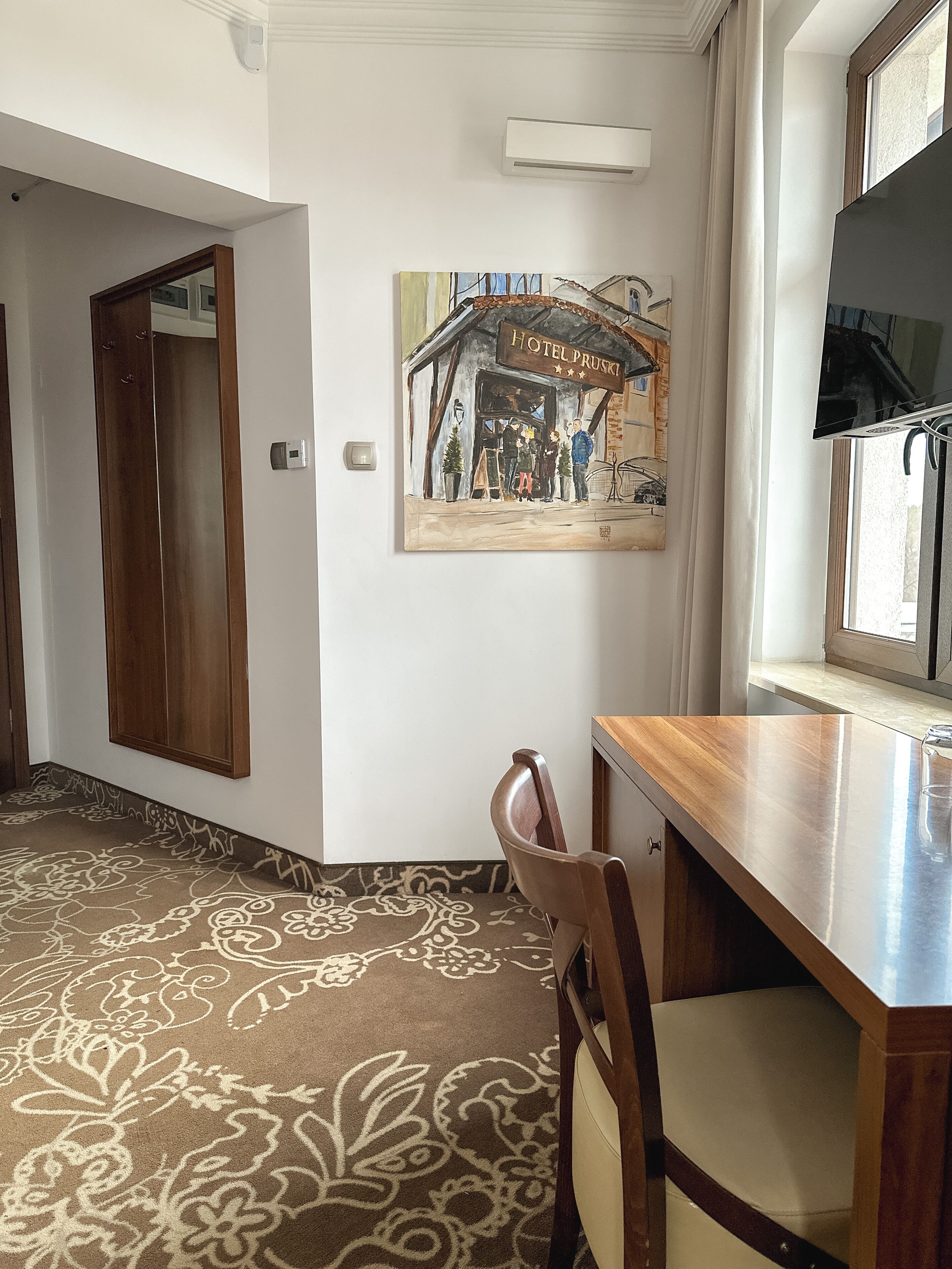 A hotel room with a wooden desk, a chair, a patterned carpet, a painting of a hotel scene, and a window with blinds.