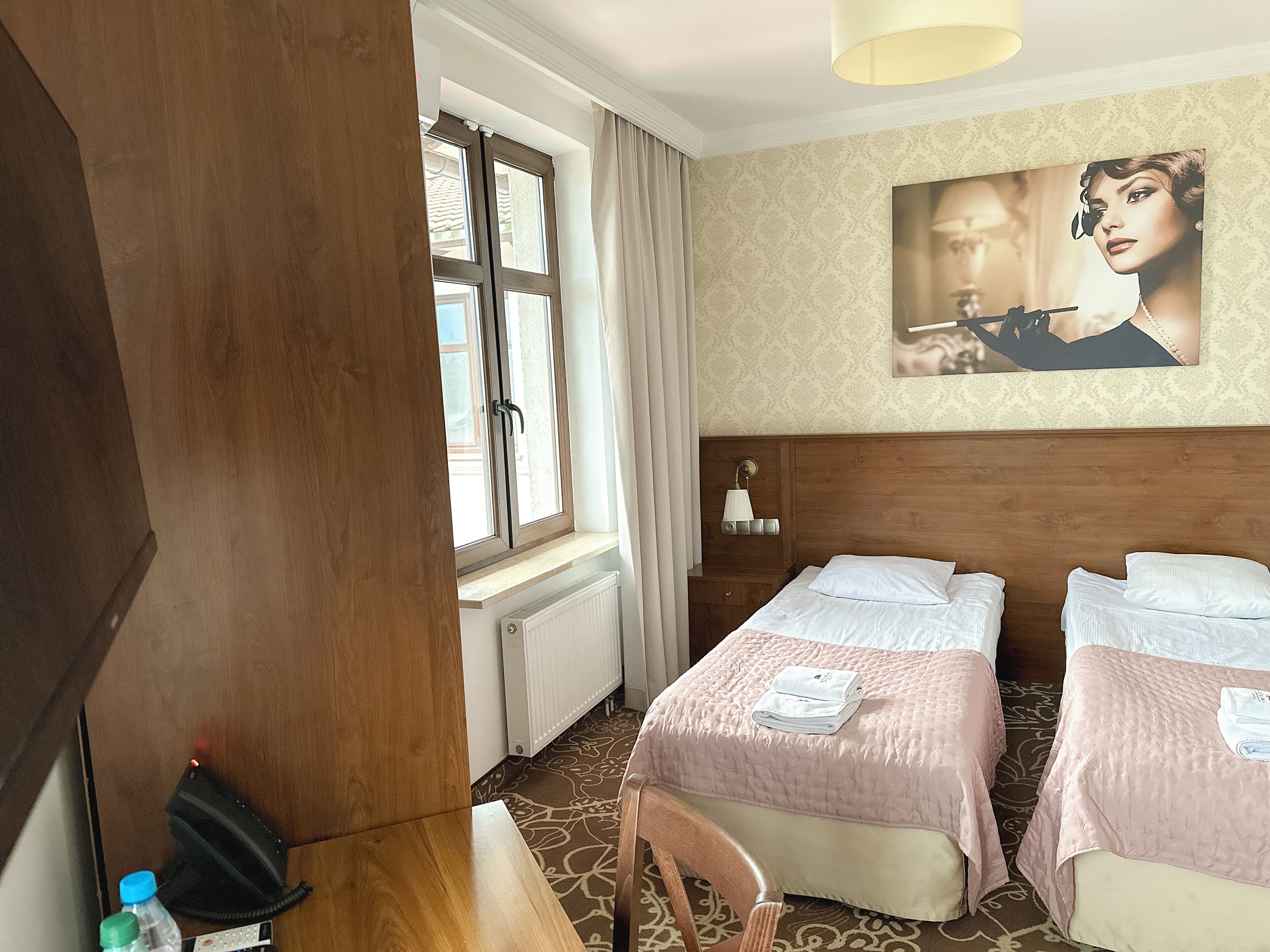 Habitación de hotel con dos camas, una ventana con cortinas, un radiador, una lámpara de pared y un cuadro.