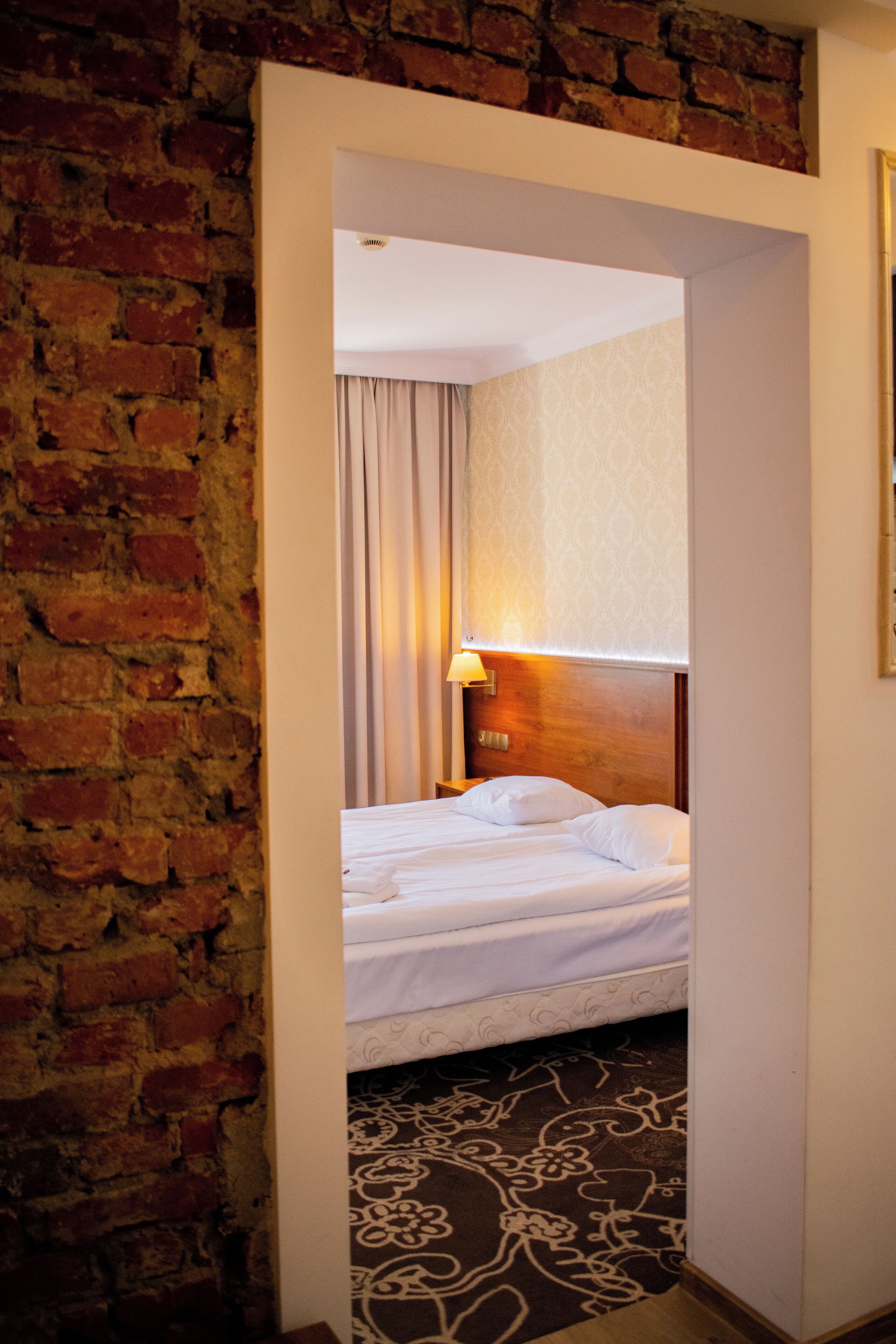 A hotel room with a brick wall, a bed with white linens, a patterned carpet, and a nightstand with a lamp.