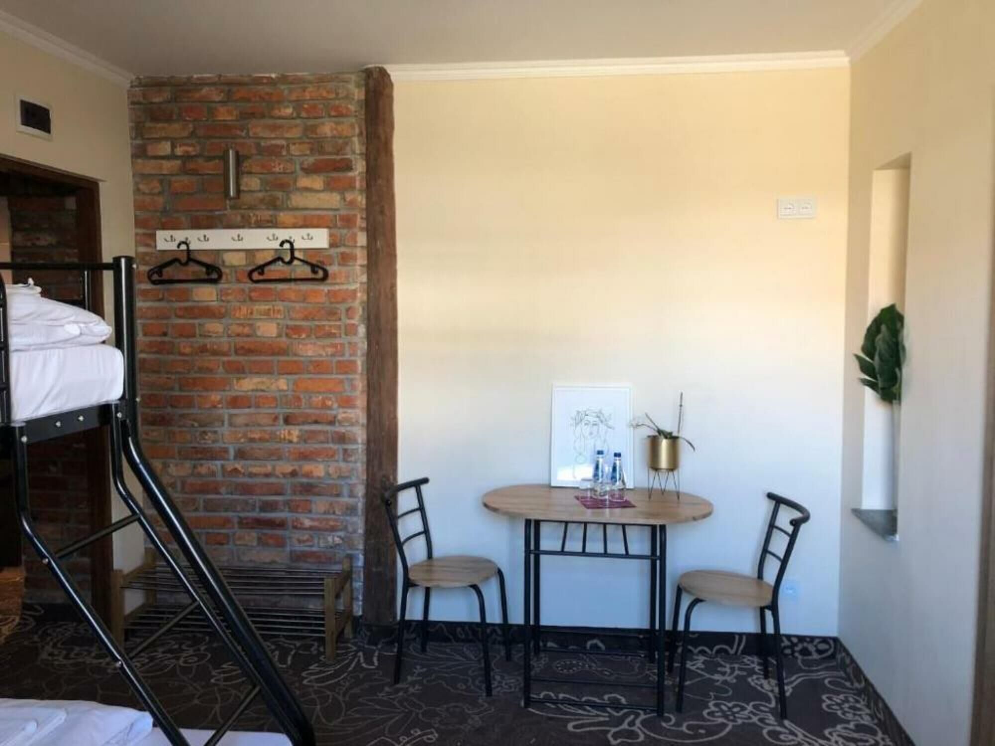 Una habitación con literas, una pared de ladrillo, una pequeña mesa con sillas, un cuadro enmarcado y una planta en maceta.