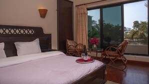 Une chambre d’hôtel avec un grand lit, deux fauteuils en osier, une petite table avec une bouteille et des verres, et une vue sur les palmiers à travers de grandes fenêtres.