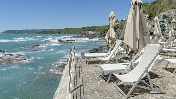 Une terrasse en bord de mer, avec des fauteuils blancs et des parasols beiges, offrant une vue sur l’océan.