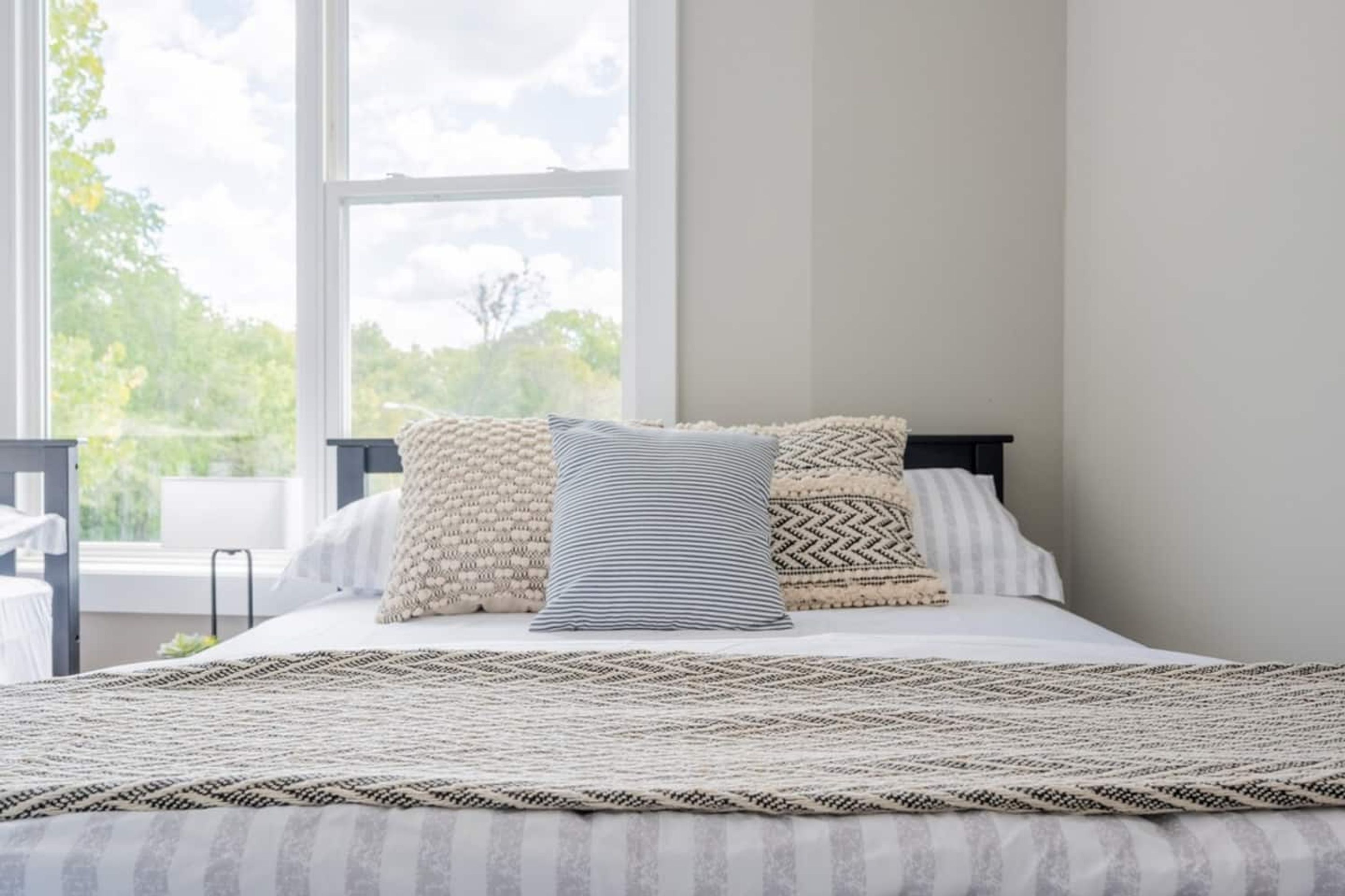 Een net opgemaakt bed met een beddekleed en kussens, een raam met uitzicht op groen, en een strak, minimalistisch interieur.