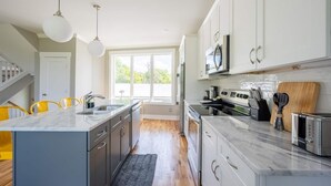 A modern kitchen with white cabinets, a marble island, and a large window providing natural light.