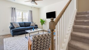A living room with a dark blue sofa, a patterned armchair, a coffee table, a ceiling fan, a staircase with a wooden handrail, and a large window with curtains. - 1 King 2 Queens Garage Yard Washer Dryer (Old Hickory)
