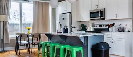 A modern kitchen with white cabinets, a stainless steel refrigerator, a microwave, and a coffee maker. There are four green stools and a black trash can.
