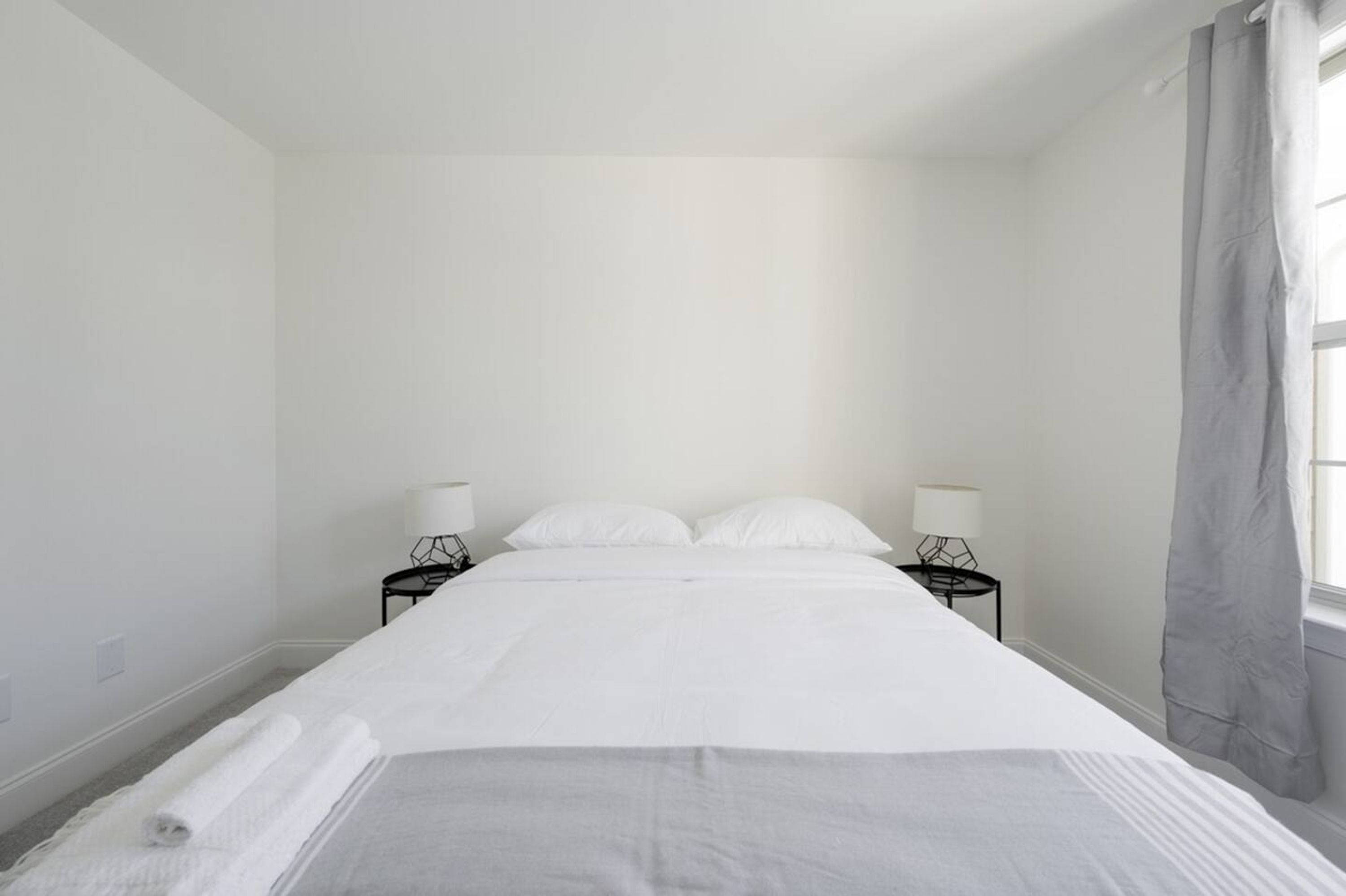 Une chambre d’hôtel au style minimaliste, dotée d’un grand lit, de deux tables de chevet avec des lampes et d’une fenêtre avec des rideaux.