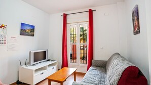 Habitación amoblada con sofá, una televisión sobre un mueble blanco, una mesita y una ventana con cortinas rojas.
