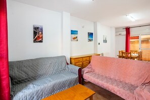 A living room with two sofas, a wooden table, and a small cabinet. There are wall-mounted pictures and a window with red curtains. - Apartment in Lepe with Pool and Garden (Lepe)