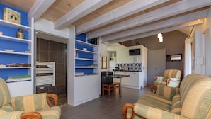 A living room with a sofa and armchairs, a kitchen area with a dining table, and a TV mounted on the wall.