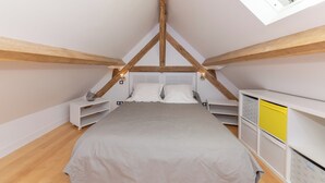 A bedroom with a sloped ceiling, wooden beams, a bed with white bedding, a white wardrobe, and a skylight.