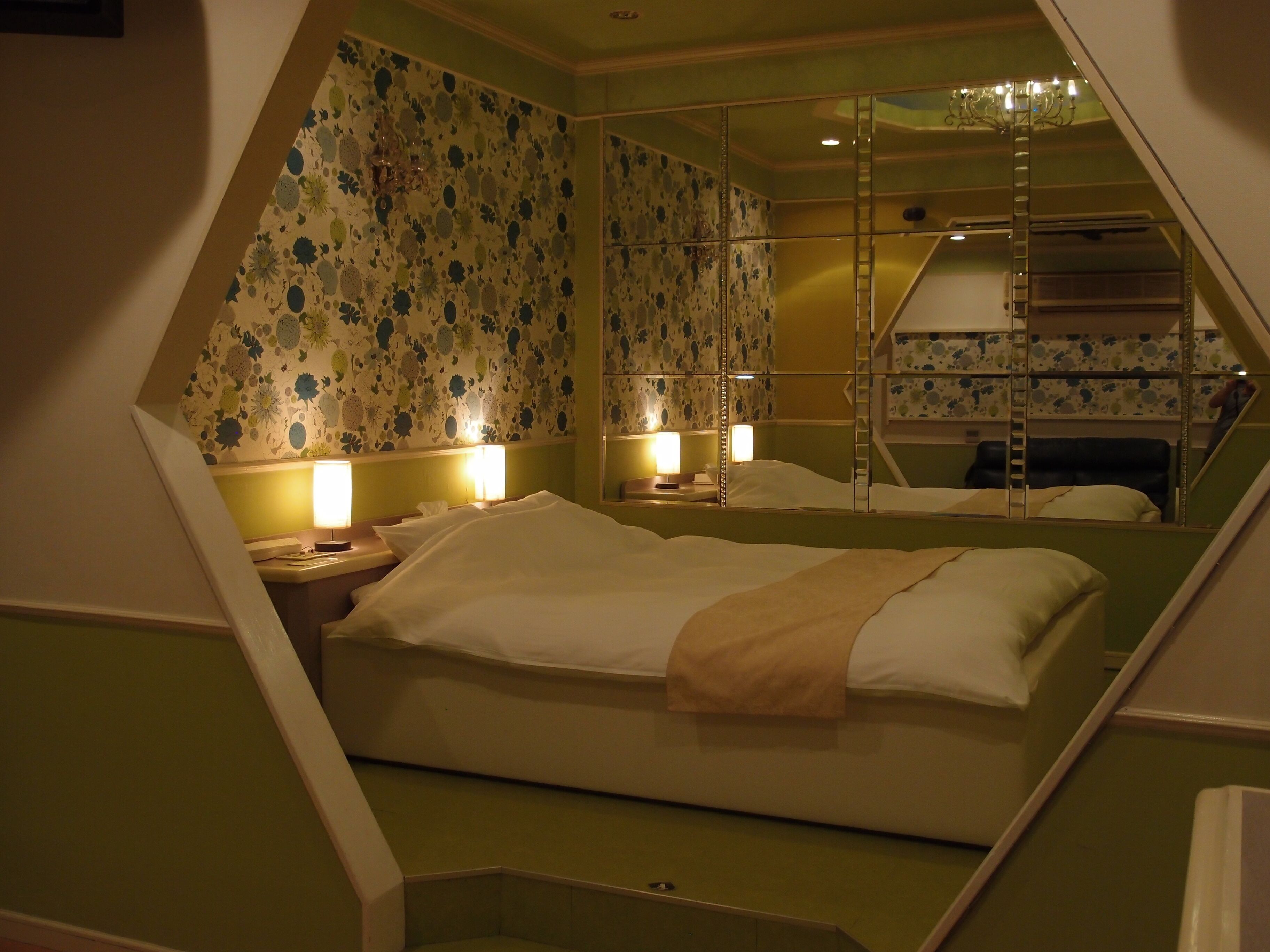 A triangular room with a bed, two bedside lamps, a mirror, and a chandelier.