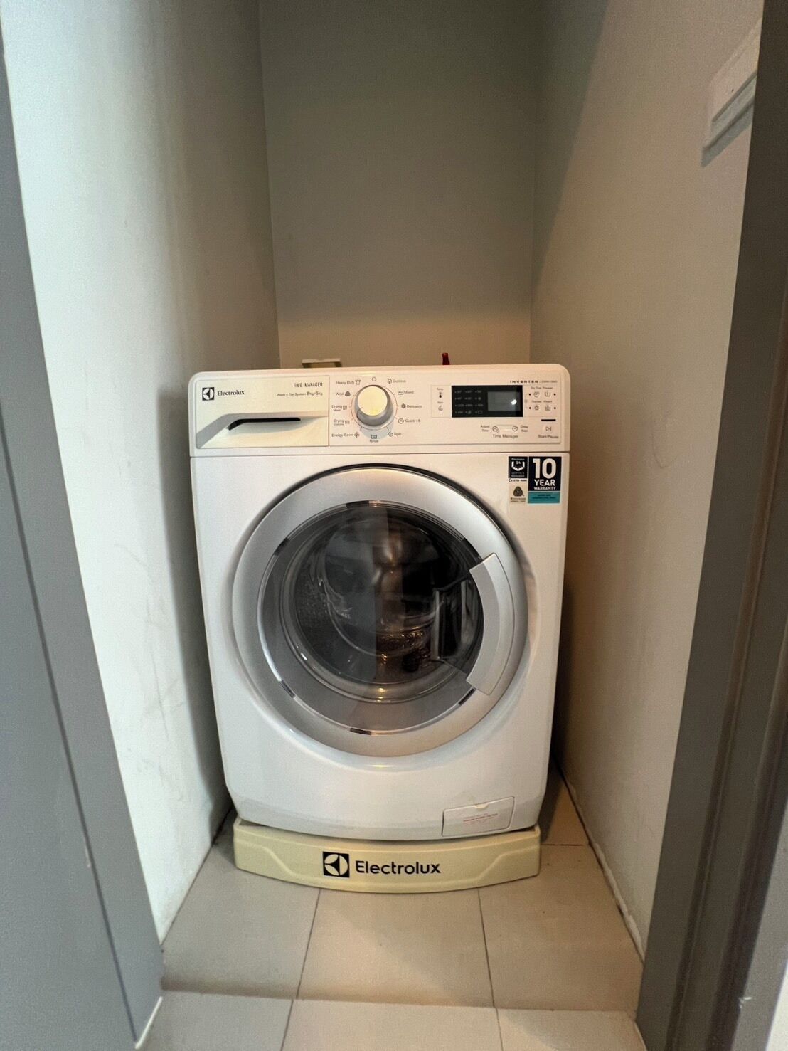 A compact, white Electrolux front-loading washing machine in a small, enclosed laundry room with a tiled floor.
