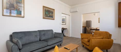 A living room with a grey sofa, a yellow armchair, and a wooden coffee table. There are two framed artworks on the wall.