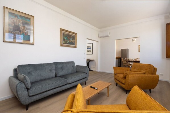 A living room with a grey sofa, a yellow armchair, and a wooden coffee table. There are two framed artworks on the wall.