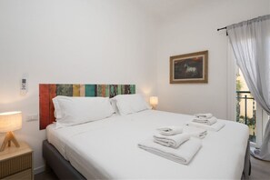 Una cama bien hecha con ropa blanca y toallas, un cuadro abstracto y colorido en la pared, una fotografía enmarco y una ventana con cortinas.