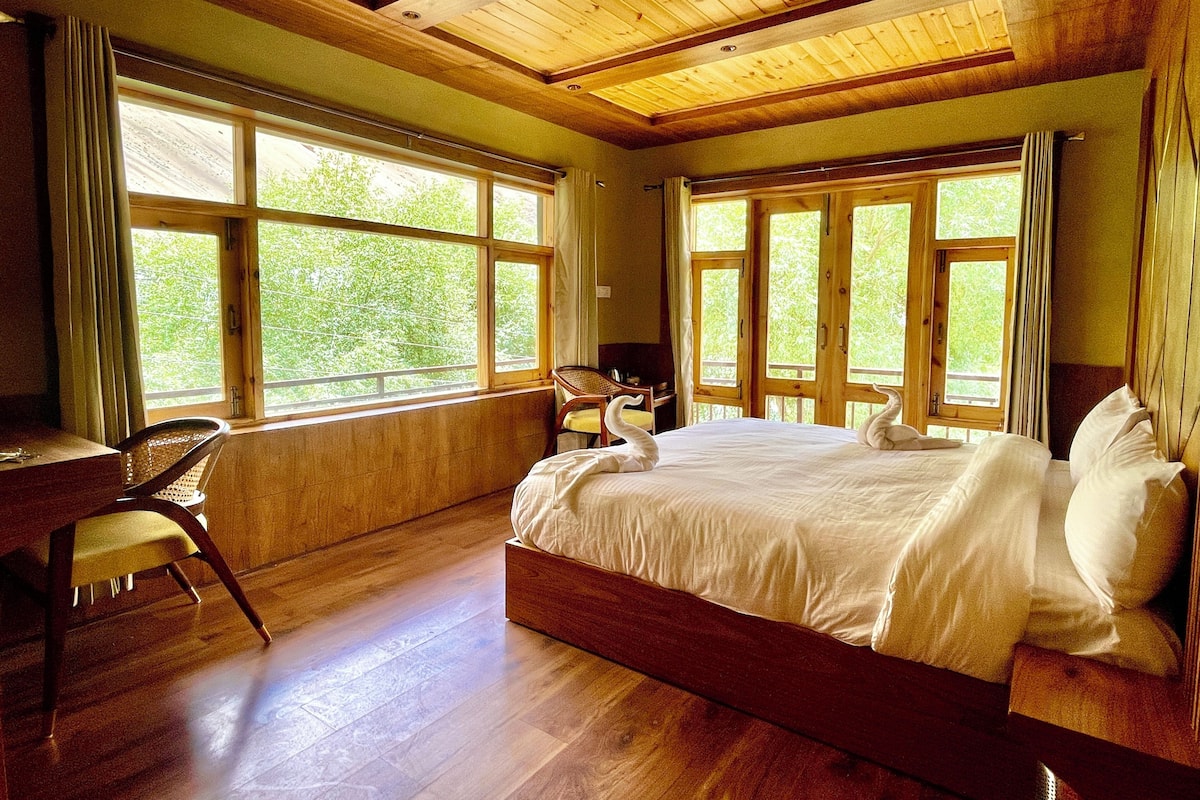 A bedroom with a large bed, wooden flooring, and multiple windows offering a view of greenery.